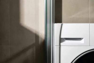Gros plan minimaliste montrant la proximité entre le coin d'une machine à laver et une paroi de douche en verre, symbolisant la cohabitation d'une douche et d'une lavatrice.