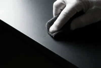 Gros plan sur une main gantée qui répare avec précision une rayure sur la surface d'un meuble laqué mat.