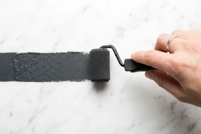 Gros plan d'un rouleau appliquant une couche de peinture grise sur du marbre blanc, symbolisant la rénovation.